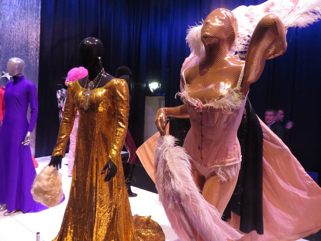 Un mannequin noir vêtu d'une robe longue à traîne dorée, un autre entièrement recouvert d'une résille dorée, dans une posture de show, vêtu d'un corset rose et tenant deux grands éventails à plume de même couleur