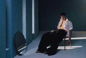 Le chanteur Omar Apollo assis chemise ouverte dans une pièce bleue devant un miroir