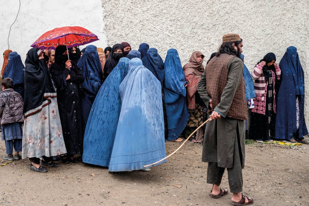 A Taliban security personnel stands guard as Afghan burqa-clad women wait in queue in the midst of a downpour to receive food supplies donated during the Islamic holy fasting month of Ramadan, in Kabul on March 25, 2025